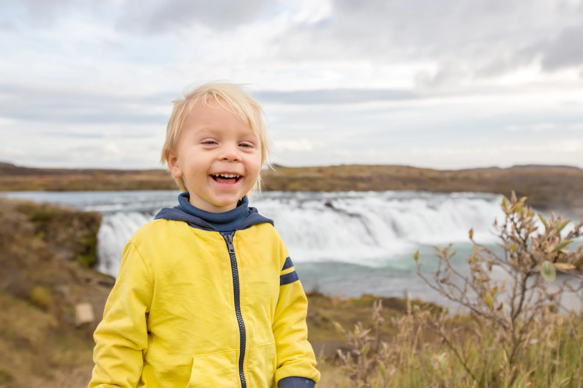A laughing young child in a yellow jacket standing near a waterfall.