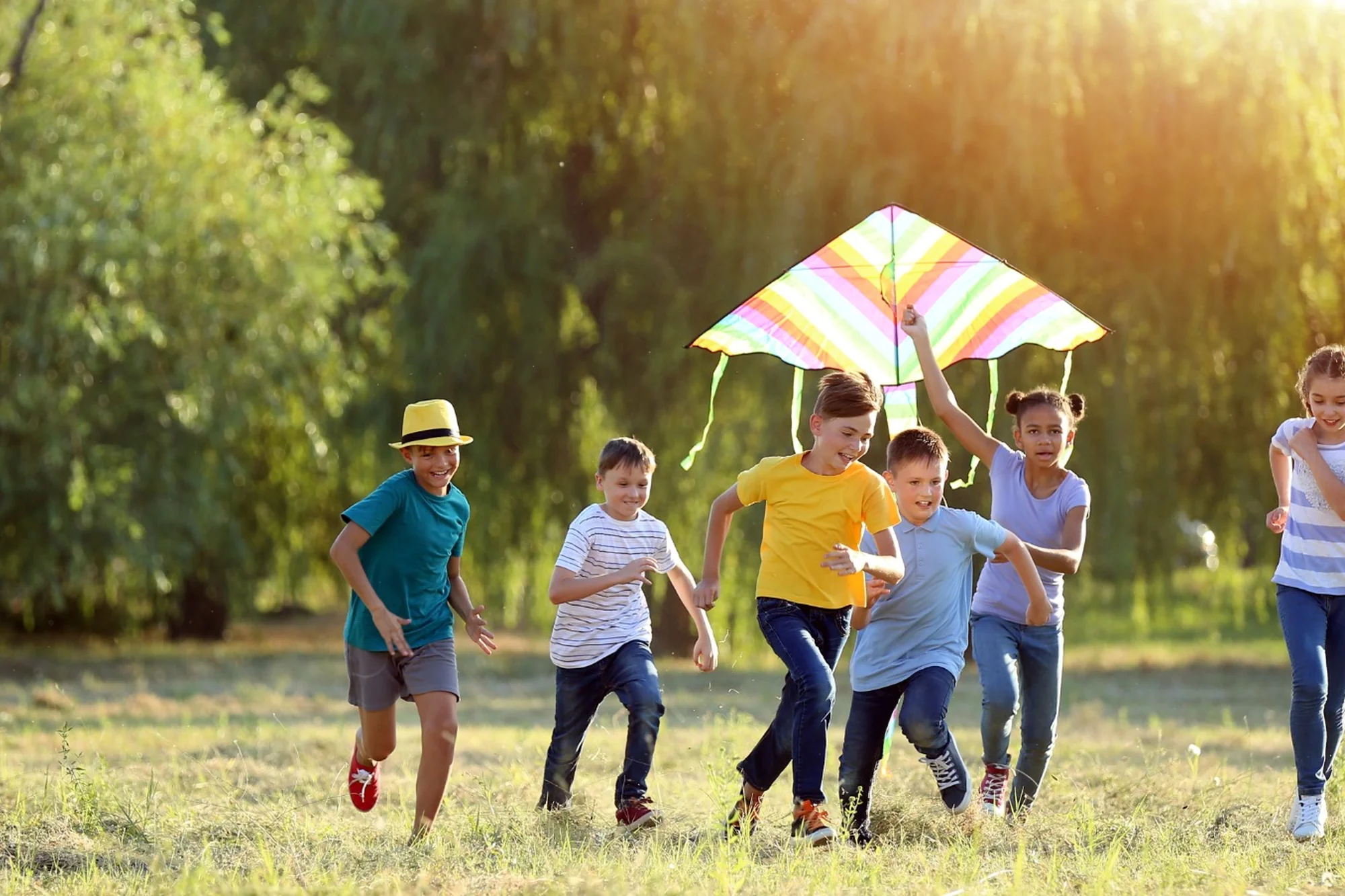 Children running through a sunlit field with a colorful kite.
