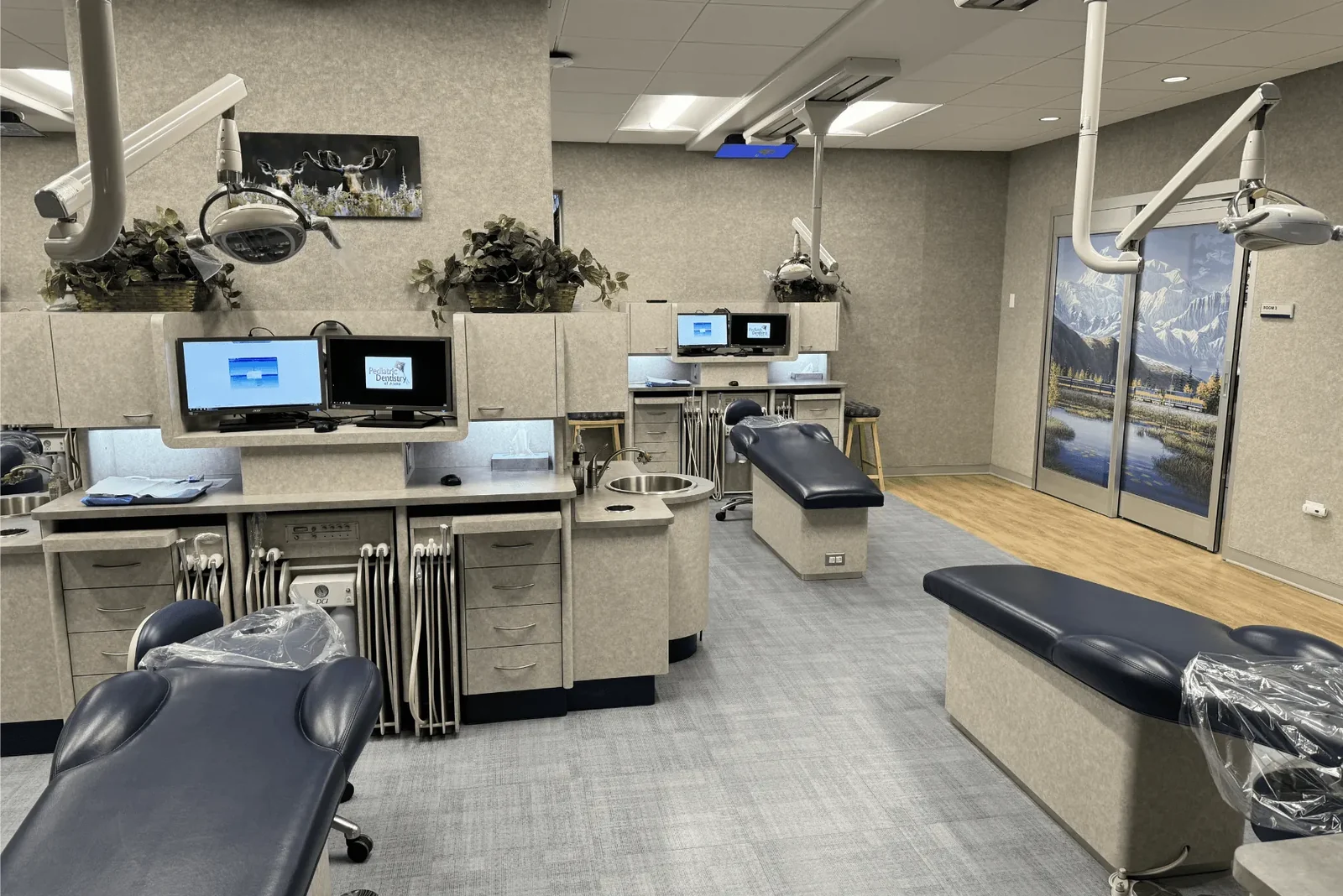 Pediatric dental treatment room with chairs, monitors, and ceiling-mounted equipment.